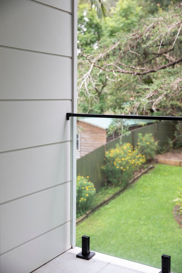 Close-up architectural view of a frameless glass balustrade or guard panel with black metal fittings, set beside a light grey horizontal-clad wall. The glazing overlooks a small green garden with lawn, planting, fencing, and neighbouring timber-clad structures.
