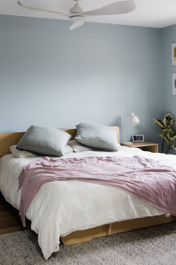 A calm, softly lit bedroom featuring a low timber bed with white bedding, pale blue-grey walls, muted grey pillows, and a textured blush-pink throw. A white ceiling fan, bedside table with a lamp and small framed item, indoor plant, wall art, and neutral textured rug complete the relaxed contemporary setting.