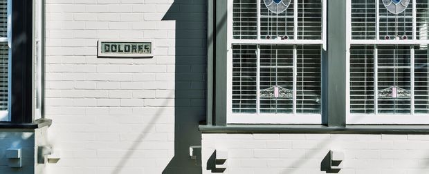 Close-up exterior view of a traditional painted brick facade with white-painted brickwork, dark green trim, and two tall multi-pane sash windows fitted with white horizontal blinds. A small plaque reading “DOLORES” is mounted on the wall, with strong sunlight casting crisp shadows across the facade.