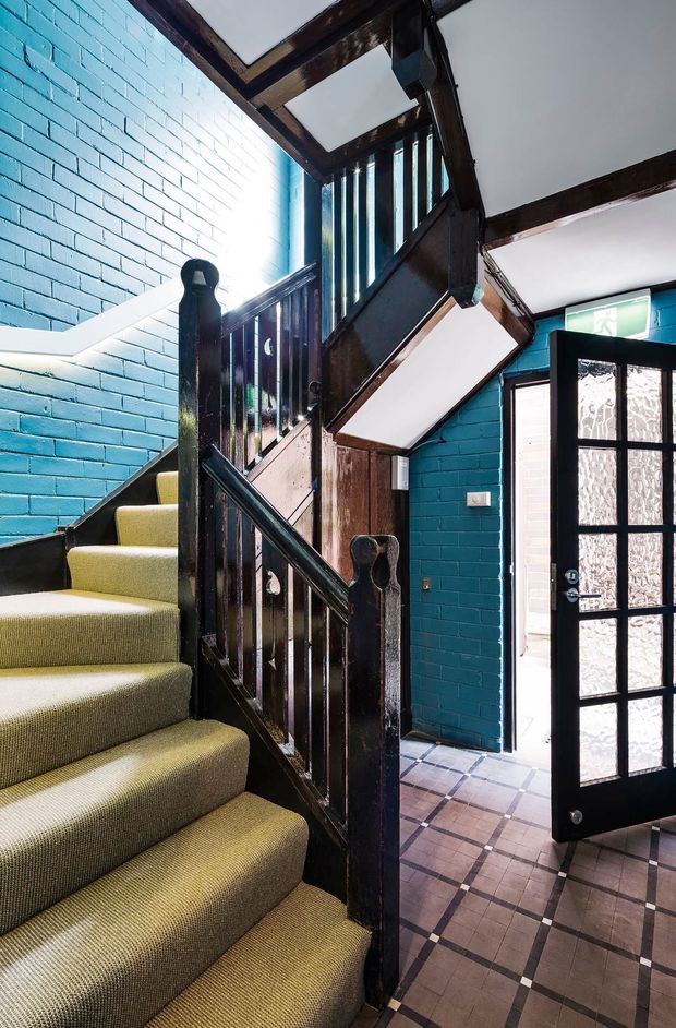 Interior view of a compact stairway and entry area. A carpeted staircase with dark stained timber balustrades rises beside vivid blue-painted brick walls. Exposed dark timber beams and stair structure frame the space, while a black glazed-panel door opens to the exterior. The floor is finished with patterned square tiles in muted brown and cream tones, creating a bold, characterful, heritage-inspired interior.