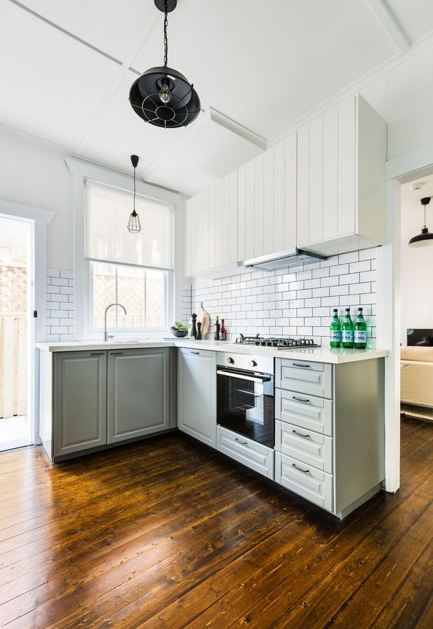 Bright, compact kitchen with white vertical-panel upper cabinetry, grey-green lower cabinets, a pale benchtop, and a white subway-tile splashback. The room features a freestanding-style stainless oven and gas cooktop, farmhouse-inspired sink area, dark polished timber flooring, and contrasting black pendant lights. A doorway reveals part of an adjacent living area.