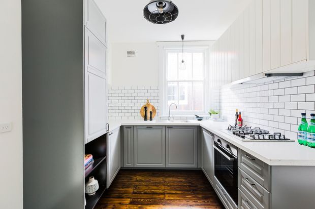 Bright, narrow galley-style kitchen with white Shaker-style upper cabinetry, dark grey lower cabinets, white subway-tile splashback, and a white benchtop. A timber-framed window above the sink provides natural light, while dark polished timber floorboards and a black ceiling light add contrast. Integrated cooking appliances and open shelving are visible along the side walls.