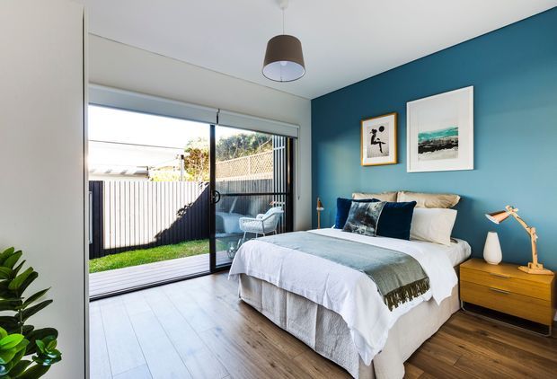 Bright contemporary bedroom with a white upholstered bed dressed in layered white and pale blue bedding, set against a deep blue feature wall. A timber bedside table with a modern lamp sits beside the bed, while large sliding glass doors open to a small fenced outdoor area. The room has pale timber flooring, white walls and ceiling, framed artwork, and abundant natural light.