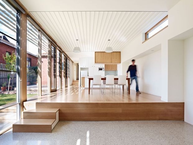 Bright open-plan interior with a kitchen and dining area positioned on a raised timber platform. Full-height timber-framed glazing runs along one side, bringing in abundant daylight and views to a neighbouring brick exterior. The space features white walls and cabinetry, timber kitchen joinery, a white vertically panelled ceiling, pendant lights, and a polished terrazzo-like floor with integrated timber steps.