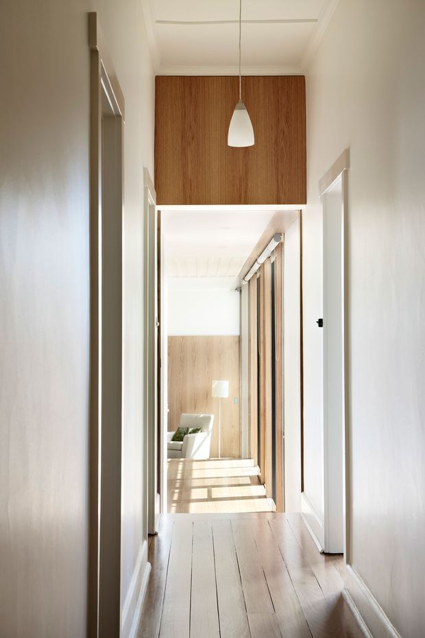 A bright, narrow residential hallway with white walls, detailed ceiling mouldings, and pale timber floorboards. A warm timber-panelled feature section sits above a wide opening at the end of the corridor, with a simple white pendant light hanging in front. The opening leads into a sunlit living area with timber panelling, glazing, and part of a sofa visible. White door frames and dark hardware create subtle contrast, while strong sunlight casts linear shadows across the floor.