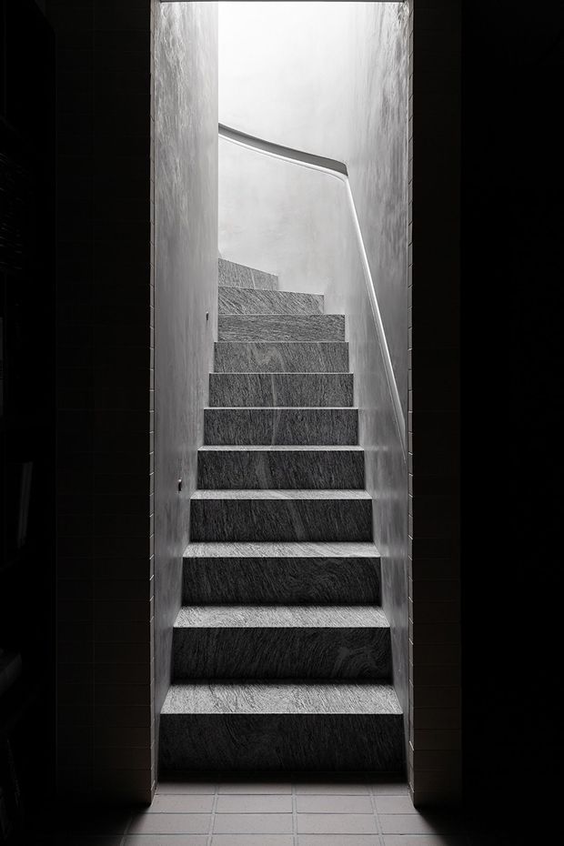 A narrow, dramatic stairway viewed from the bottom, with dark textured stone or tile-clad steps ascending through a tall opening. Smooth light-grey walls and a slim metal handrail contrast with the deep shadowed side walls, while a tiled floor is visible at the base.