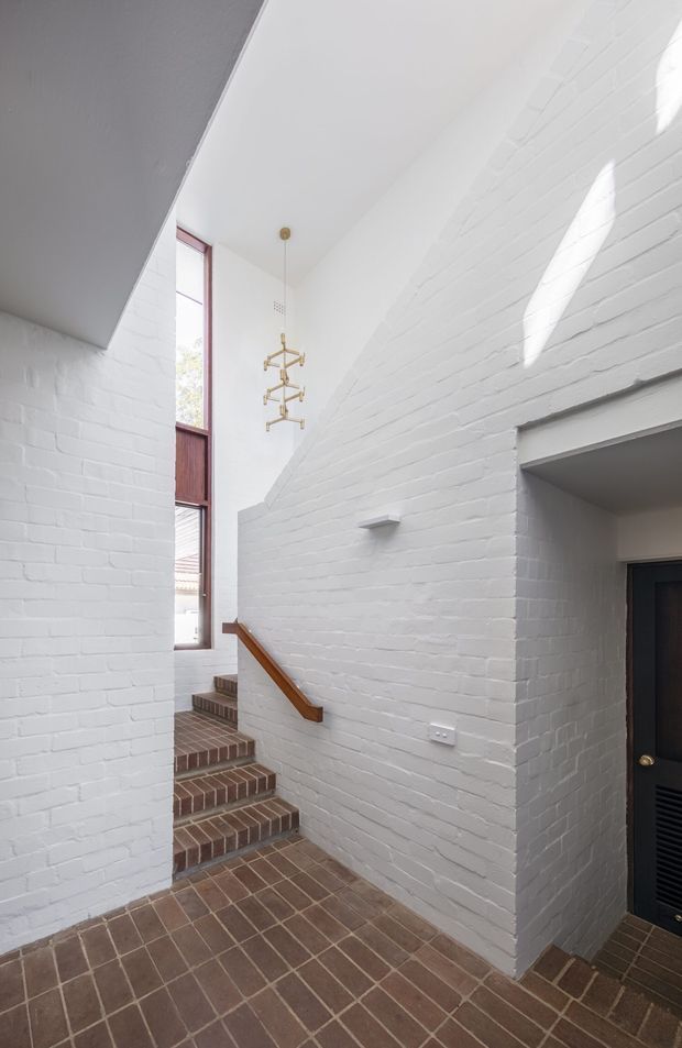 Bright narrow interior stair hall with white-painted exposed brick walls, a brick-tiled floor and staircase, tall vertical glazing, a timber handrail, and a sculptural brass pendant light. A dark louvred door opens off the hall. The space has strong daylight and a simple loft-like character.
