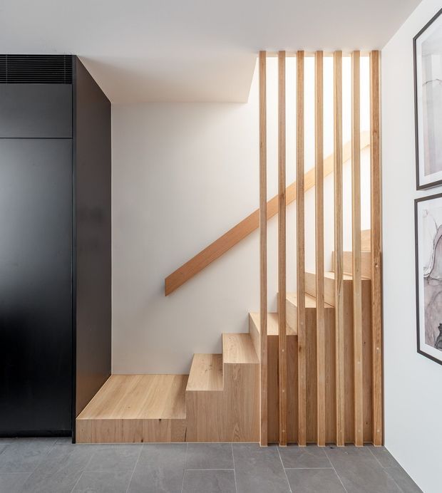 A contemporary interior stairway with pale timber-clad steps and matching handrail, screened by evenly spaced vertical timber battens. The stair is adjacent to a tall dark built-in cabinet, with white walls, framed artwork, and large grey floor tiles. The partially enclosed stair design creates a clean, warm, minimalist architectural composition.