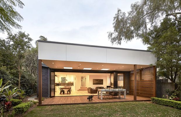 A contemporary single-storey pavilion-style house extension viewed from the garden. The open-sided structure has a flat roof with a broad white fascia, dark brick side walls, timber soffit, timber decking, and a horizontal timber louvred privacy screen. Large glazed openings connect the covered outdoor dining area with a warmly lit interior living and dining space. Mature landscaping surrounds the building.