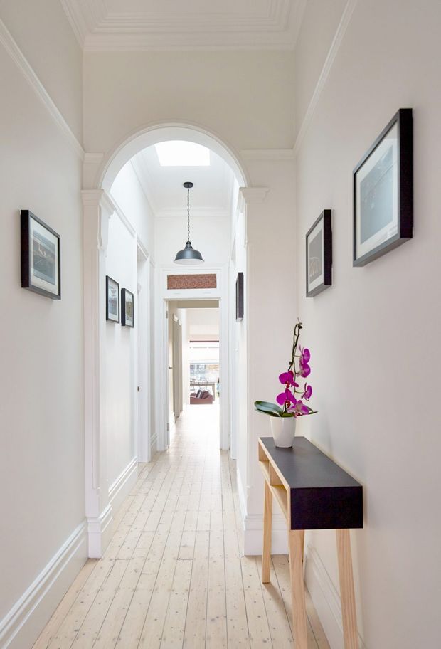 Bright, elegant residential hallway with white walls, detailed moulded archways and corniced ceiling, framed artwork, a pendant light, and a slim console table with an orchid. The corridor extends toward a brighter living area, with pale painted timber floorboards creating an airy, refined feel.