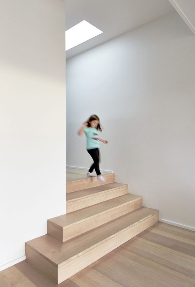 Bright minimalist interior view of a sculptural timber stairway with broad open box steps and matching timber flooring. White walls and ceiling create a clean, airy setting, with a rectangular skylight bringing in natural light. A child is moving down the stairs.