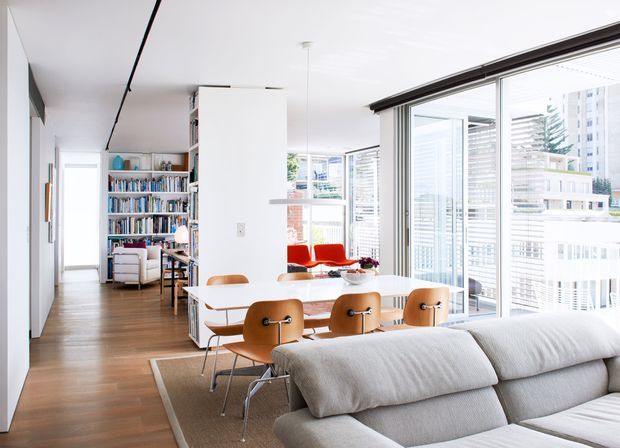 Bright contemporary open-plan apartment interior with a dining area in the foreground, a large grey sectional sofa, white walls, warm timber flooring, extensive floor-to-ceiling glazing, and a compact living or reading area with built-in bookshelves beyond. The space has strong urban views and abundant natural light.