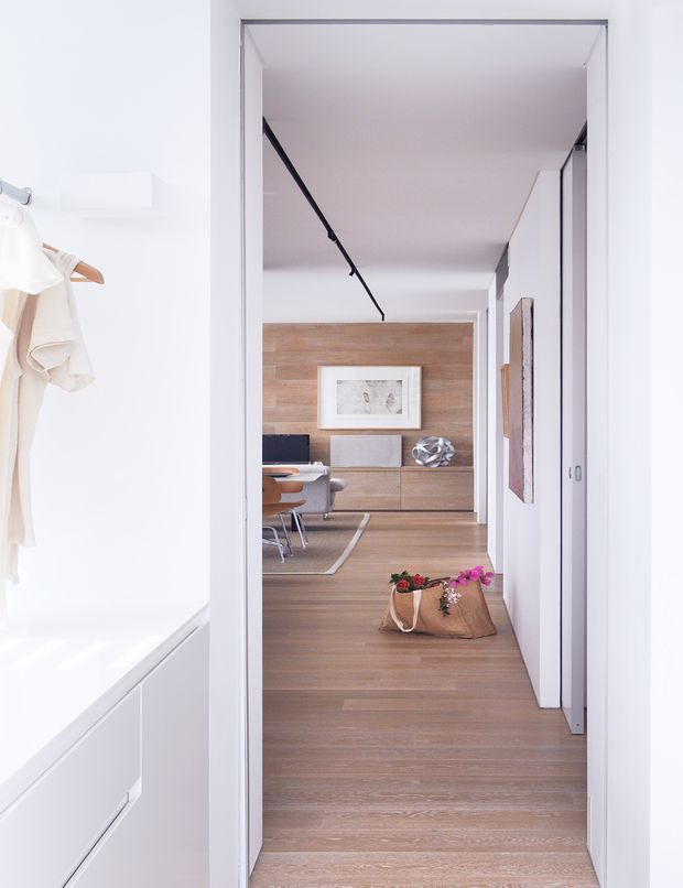A bright, minimalist interior view looking through a hallway toward a dining and living area. The foreground includes a white built-in bath or cabinetry zone and a small clothing rail, while the central space has warm timber flooring, a timber feature wall with low built-in cabinetry, a dining table and chairs, framed artwork, and white floor-to-ceiling joinery. A black linear track light runs along the ceiling, creating a clean contemporary composition.