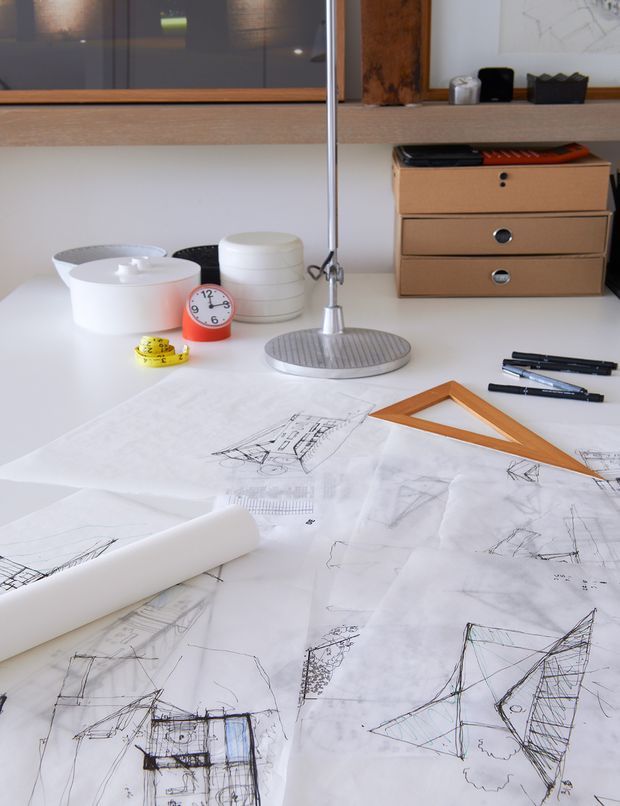 A close-up view of a drafting workspace with architectural sketches and tracing paper spread across a white desk. The desk holds a rolled drawing sheet, orange triangular ruler, pens, tape measure, desk lamp, small clock, white containers, and wooden document drawers. A wall-mounted shelf and framed artwork are visible in the background. The scene has a clean, creative studio atmosphere with restrained mid-century-inspired furnishings.