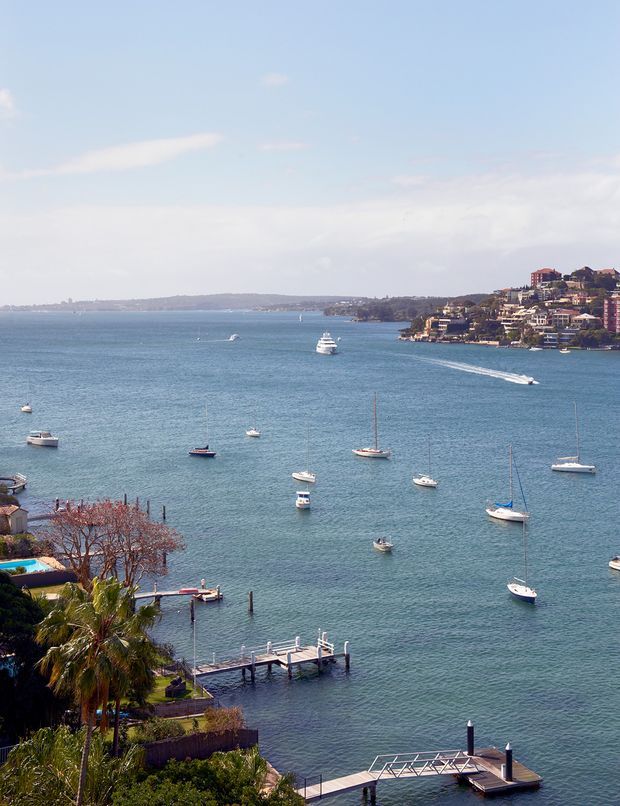 A wide elevated view across a sheltered coastal harbour, with numerous anchored sailboats and motorboats, wakes from passing vessels, and several private jetties extending into the water. Residential buildings line the opposite wooded shoreline and rise up a hillside, while landscaped waterfront properties with palm trees and docks occupy the foreground. The scene is bright and scenic, dominated by blue water and sky.