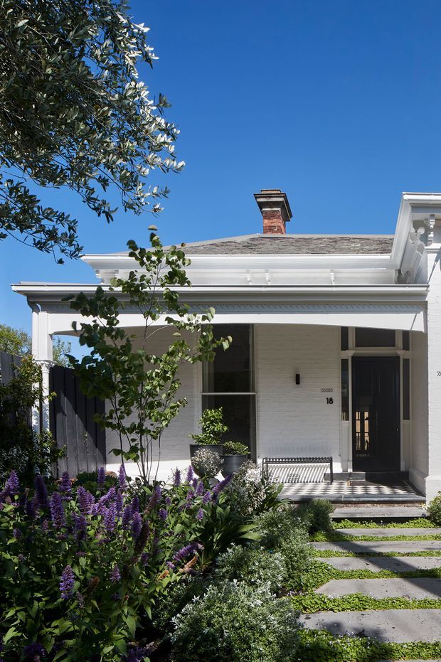Front exterior view of a refined white-painted brick house with a shallow hipped roof, prominent decorative cornice and porch framing, a brick chimney, dark entry door, and landscaped garden. A stepping-stone path leads through dense flowering and ornamental planting to the raised entrance. The facade has traditional detailing and a bright, well-maintained appearance.