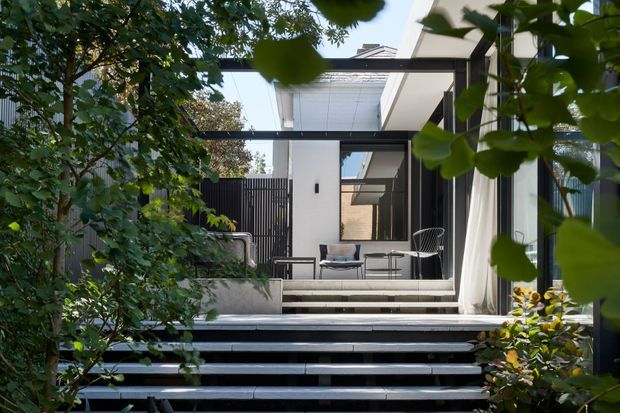 A modern outdoor terrace or patio viewed through lush garden planting, with broad dark steps leading up to a raised entertaining area. The space features black steel framing, white walls, large glazed openings with sheer curtains, a partially covered roof, and compact outdoor seating. The composition highlights the connection between contemporary architecture and landscaped surroundings.