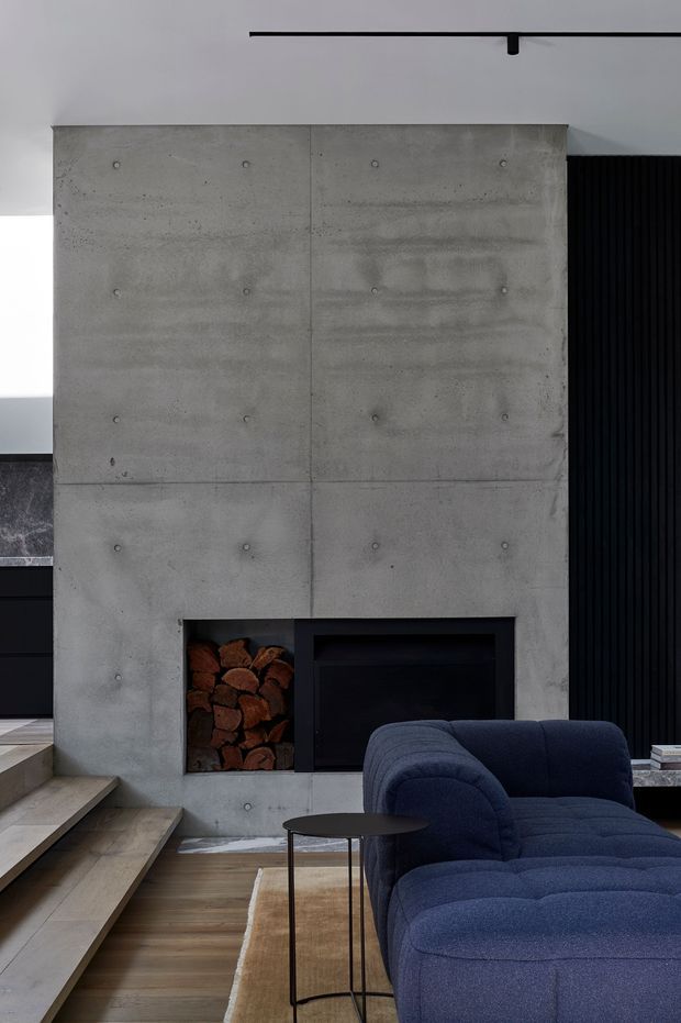 A moody contemporary living room centred on a large exposed concrete fireplace wall with visible formwork joints and bolt marks. A dark recessed fireplace and built-in firewood niche sit below, beside a black vertical slatted feature wall. Timber flooring, broad timber stair treads, a deep navy sectional sofa, and a slim black side table complete the space.