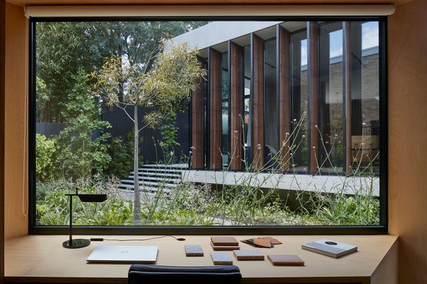Interior view from a timber-lined study or workspace looking through a large picture window toward a contemporary building. The foreground has a built-in timber desk, task lamp, notebooks and samples, while outside is a landscaped garden and a modern facade with tall vertical timber-framed glazing, dark cladding and a projecting flat roof.