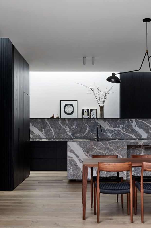 A refined open-plan kitchen and dining area with full-height dark timber cabinetry, a dramatic veined stone island and matching backsplash, and a warm timber dining table with mid-century style chairs. The room has a pale timber floor, white ceiling and wall surfaces, black architectural lighting, and restrained decorative styling.