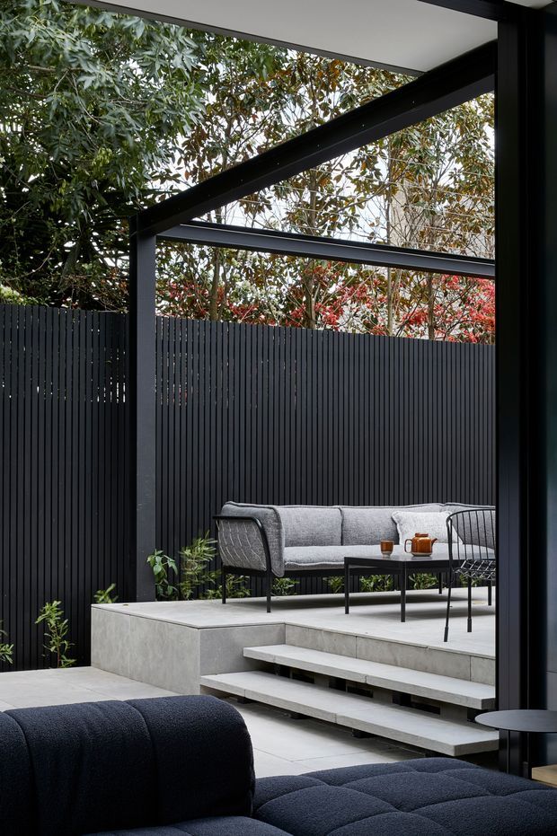 A refined covered outdoor lounge or courtyard framed by a black-painted steel structure with exposed diagonal beams. The space features a dark vertical-slat privacy fence, pale concrete stepped platforms and built-in seating, a grey outdoor sofa, black metal chairs and tables, and dense planting visible beyond the enclosure. A dark upholstered sofa occupies the foreground, creating a layered indoor-outdoor composition. The palette is predominantly charcoal, grey and concrete tones with natural green foliage and muted red foliage accents.