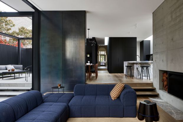 Open-plan contemporary living area with a large deep-blue sectional sofa in the foreground, facing a concrete fireplace with integrated wood storage. Blackened metal or dark panelled walls divide the living room from a dining area and compact kitchen, while full-height glazing opens to an outdoor terrace. The kitchen includes dark cabinetry, a pale stone island with bar stools, and a timber dining table beyond. The space has a refined, high-contrast material palette and strong indoor-outdoor connection.