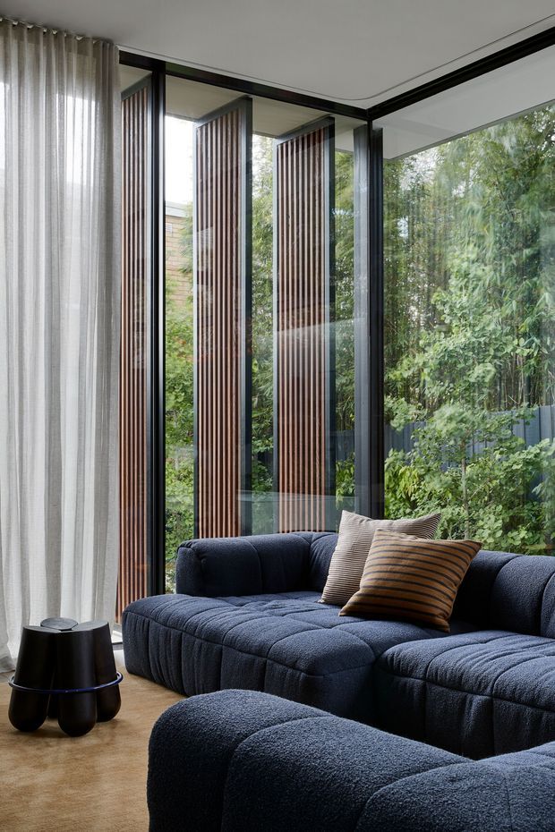A sophisticated living room with a deep navy modular sectional sofa and matching ottoman arranged beside full-height glazed corner windows. Sheer floor-to-ceiling curtains soften the light, while dark-framed glazing and vertical timber screening create a strong architectural backdrop. A small black sculptural side table and striped neutral cushions complete the refined, contemporary setting. Dense greenery is visible outside.
