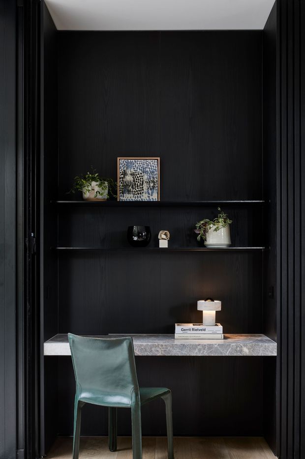 A compact, dark-toned built-in workspace set into a wall niche. Black timber-look wall panelling and shelving frame a pale stone desk surface, with a green upholstered chair, small table lamp, plants, artwork, and decorative objects. Vertical black slatted detailing borders both sides, while a light angled ceiling plane adds contrast. The overall composition is refined, moody, and highly tailored.