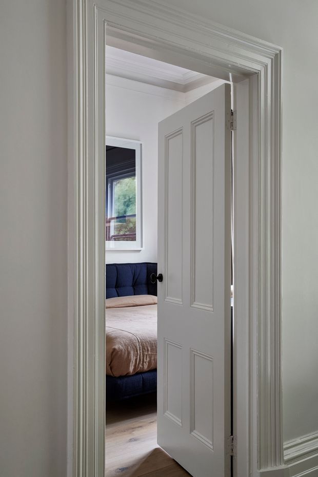 View through a partially open, white paneled interior door with ornate layered trim into a bedroom. The bedroom contains a dark navy upholstered bed with a pale beige duvet, light timber flooring, a window, and white walls with decorative ceiling and wall mouldings. The foreground is primarily an interior hallway or doorway, with the bedroom visible beyond.