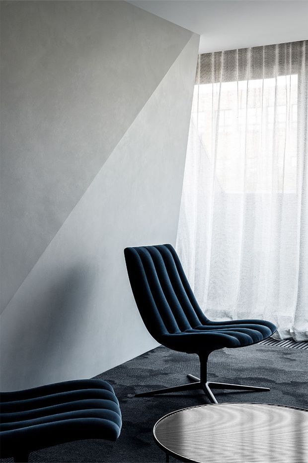 A minimalist interior corner featuring a sculptural black upholstered lounge chair with channel tufting and a matching partial ottoman, positioned on dark textured carpet beside a round reflective coffee table. Pale sculpted walls with sharp geometric planes and sheer floor-to-ceiling curtains create a quiet, monochromatic setting.