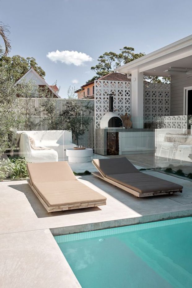A bright contemporary backyard pool area with turquoise water, pale concrete paving, two timber-framed sun loungers, glass pool fencing, and a covered outdoor entertaining zone. A white breeze-block privacy wall incorporates a curved outdoor fireplace, with glimpses of a light-filled interior and surrounding residential homes beyond.