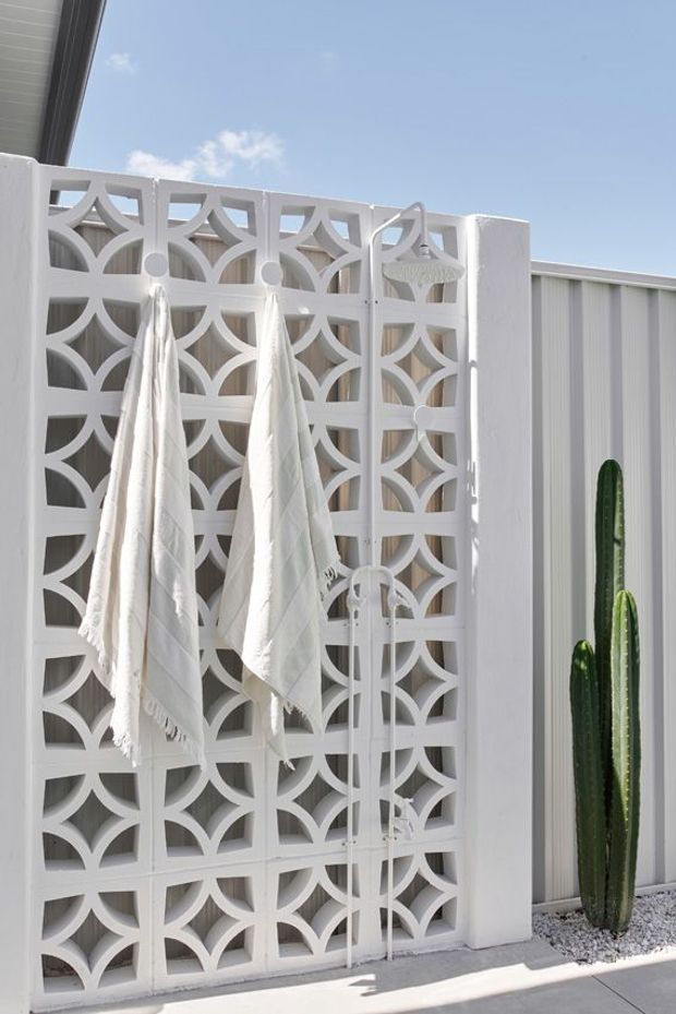 Bright exterior courtyard or patio detail featuring a white decorative breeze-block screen with repeating geometric cut-outs. White towels hang from the screen beside an outdoor shower, with a tall cactus in a gravel bed and corrugated light-grey wall cladding in the background. The scene has a clean, sunlit, resort-like character.