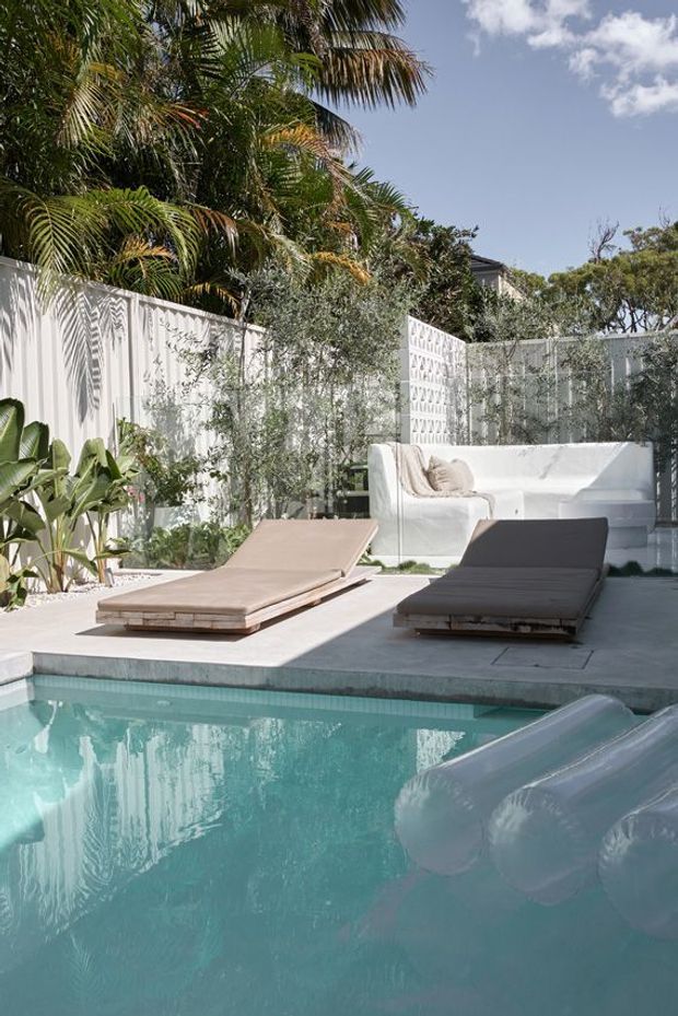 A bright landscaped outdoor pool area with turquoise water in the foreground, a pale concrete pool deck, two low timber sun loungers, and a white built-in outdoor sofa. Tropical and Mediterranean-style planting is enclosed by a decorative white breeze-block privacy wall, creating a private resort-like courtyard.