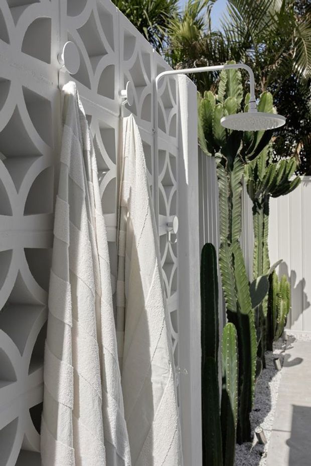 A bright outdoor shower area with a white rainfall shower fixture mounted against a decorative geometric breeze-block wall. White towels hang from integrated hooks, while tall sculptural cacti and tropical foliage line the adjacent white-fenced walkway. The scene has a crisp, sunlit resort-like appearance with strong geometric shadows.