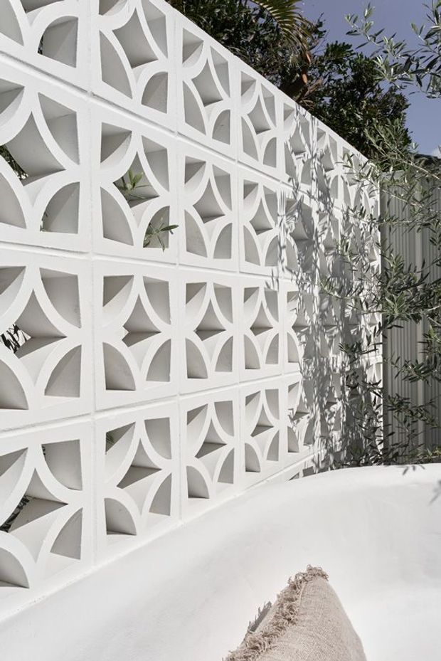A sunlit exterior courtyard or terrace featuring a white decorative breeze-block screen wall with repeated geometric cut-out patterns. White rendered surfaces, soft shadows, and olive-like planting create a bright, Mediterranean-inspired outdoor setting.