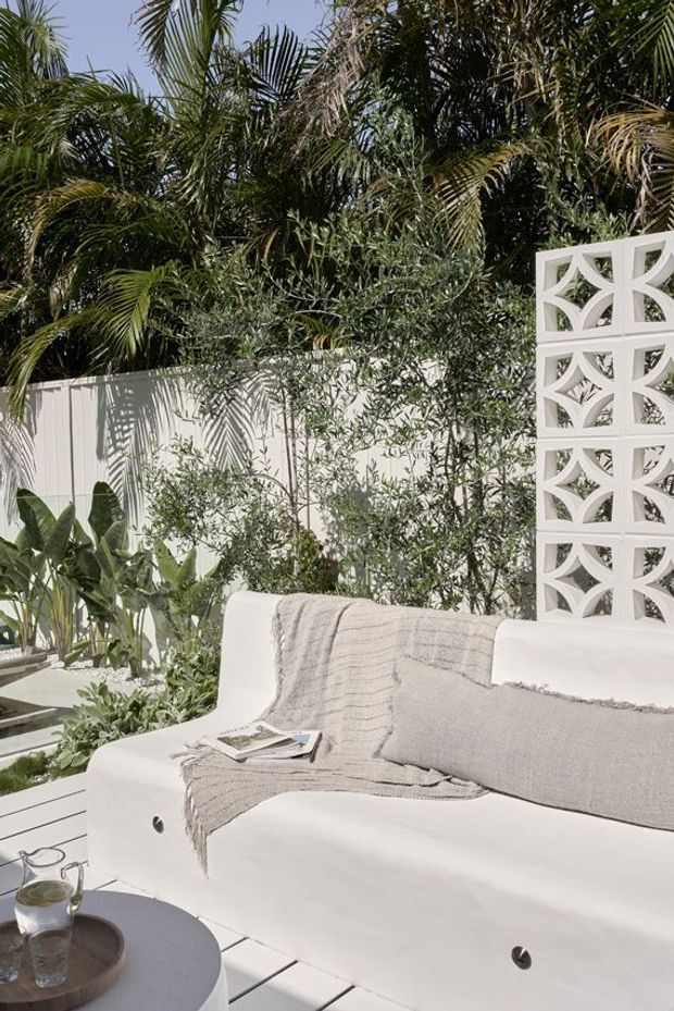 A bright tropical outdoor lounge area with a white built-in or modular sofa, neutral textured throw and cushions, and a small round coffee table. The space is enclosed by a white decorative breeze-block screen and fencing, surrounded by dense palms and lush planting, creating a private resort-like courtyard setting.