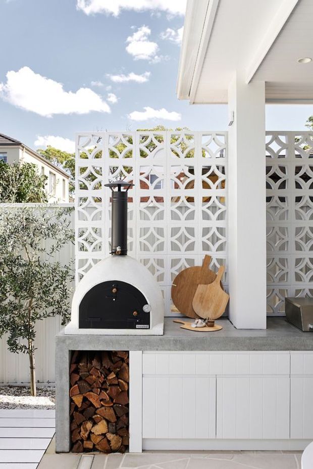 A bright outdoor entertaining area with a built-in wood-fired pizza oven, concrete benchtop, white tiled cabinetry, stacked firewood storage, and decorative white breeze-block screening. Timber serving boards and a potted olive tree add natural accents beneath a covered roofed structure.