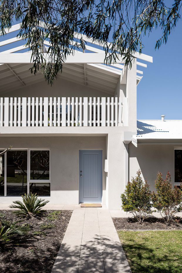 Exterior view of a bright, white two-storey residential home. The facade features a covered upper balcony with vertical white privacy battens, a broad sloping roof canopy, pale blue-grey front door, large windows, and a concrete path leading through landscaped garden beds. The building has crisp contemporary lines and a light coastal character, with a corrugated metal roof visible on the adjoining single-storey section.