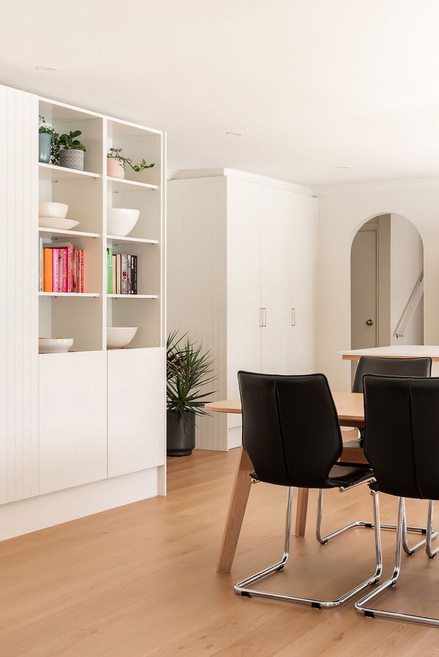 Bright contemporary dining area with a light timber floor, white built-in display shelving and cabinetry, potted plants, books and ceramics. A timber dining table with black cantilever chairs occupies the foreground, while an arched opening reveals a stairway beyond. The room has a clean, warm, minimalist interior with a low, white ceiling and integrated storage.