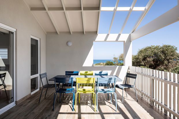 A bright covered outdoor terrace or balcony furnished with a colourful outdoor dining set. The space has timber-look flooring, white-painted walls and exposed roof beams, a translucent roof section and open sides with white vertical balustrades. Ocean and vegetation are visible beyond the terrace, while strong sunlight creates geometric shadows across the floor and furniture.
