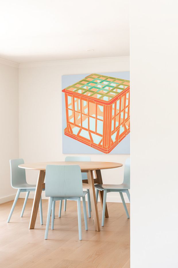 Bright, minimal dining area viewed through a partial wall opening, with a round light-wood dining table surrounded by pale blue upholstered chairs. The room has white walls and ceiling with simple cornice detailing, light timber flooring, and a colourful geometric artwork featuring an architectural cube motif.