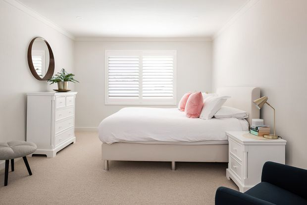 Bright, calm bedroom with a neatly made upholstered bed, white bedding and pink accent cushions. The room has pale neutral walls, beige carpet, white plantation shutters, white traditional-style drawer chests, an oval wall mirror, a small plant, and a floor lamp. A dark upholstered armchair is partially visible in the foreground.