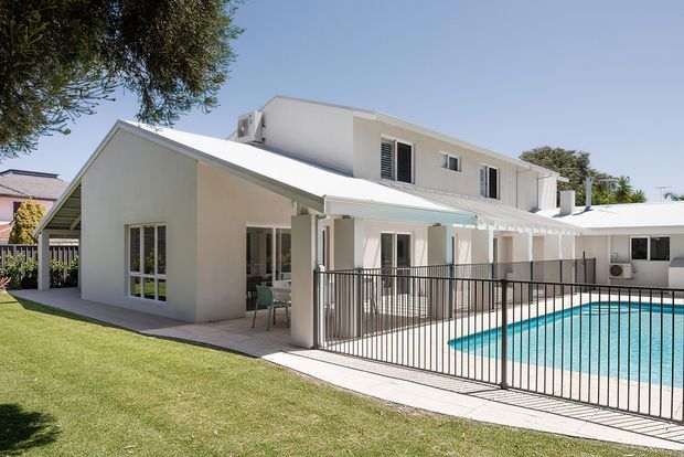 Exterior view of a bright white multi-level residence surrounding a swimming pool. The home combines a steeply pitched front gable with a larger flat-roofed upper volume, covered outdoor areas, glazed doors and windows, and a black metal pool safety fence. A manicured lawn and mature planting surround the house.