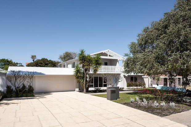 A bright exterior view of a substantial white two-storey residence, featuring a broad paved driveway, attached double garage, covered upper-level balcony, white balustrades, and a prominent pitched gable roof. Landscaped garden beds and mature trees frame the facade.