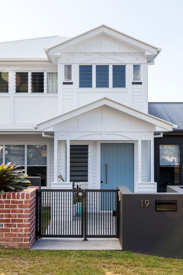 Front exterior view of a compact multi-storey residence with a white weatherboard facade, pitched gable roofs, decorative gable detailing, horizontal louvred windows, and a pale blue entry door. A black metal pedestrian gate and dark rendered front wall sit beside a red brick planter and landscaped front boundary.