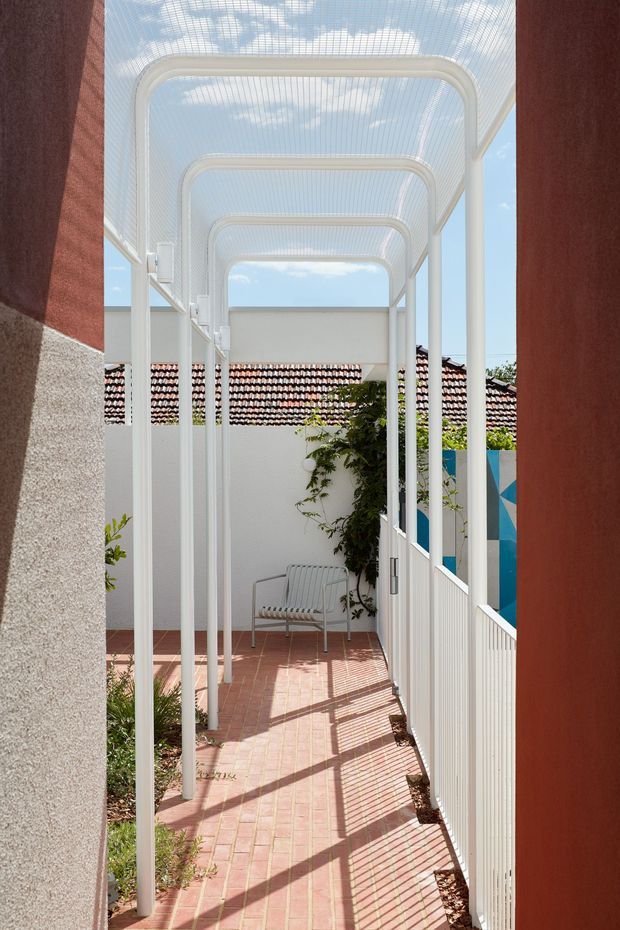 A narrow, sunlit covered outdoor walkway or courtyard passage framed by red and pale textured walls. Repeating white metal arches support a translucent mesh canopy, casting strong linear shadows across a pink terracotta-paver floor. A white metal bench, climbing greenery, garden planting, and a colourful geometric privacy screen create a bright contemporary courtyard setting.