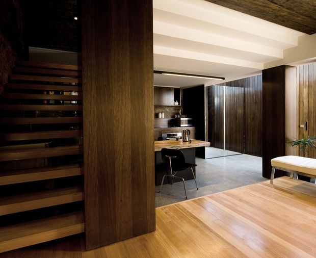 A refined open-plan interior combining a warm timber dining area with a compact contemporary kitchen. A dark timber open-riser staircase and full-height timber-clad partition frame the space, while the kitchen features dark cabinetry, a timber island benchtop, stainless steel appliances, and a pale stone or concrete-look floor. White exposed ceiling beams contrast with the darker wood finishes, creating a warm, sophisticated and architecturally detailed interior.