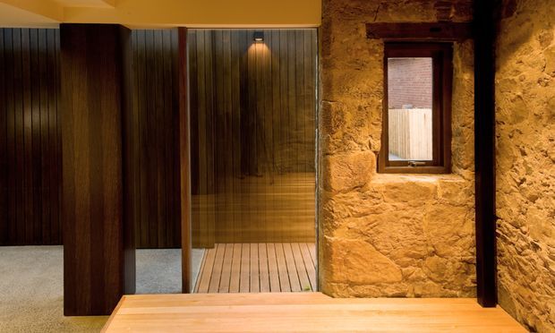 Warmly lit interior featuring a compact timber-lined wet area or sauna-like room behind glass, set within a rugged exposed stone wall. Dark vertical timber panels, a timber slatted floor, black-framed glazing, and a small high-set window create a rustic yet refined atmosphere.