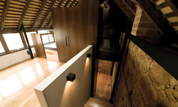Interior view of a dramatic split-level circulation space with a timber-lined upper room, exposed pitched roof structure, polished light timber flooring, and a stair descending beside a rough masonry feature wall. Dark timber beams, black metal details, and concealed wall lighting create a warm, moody architectural atmosphere.