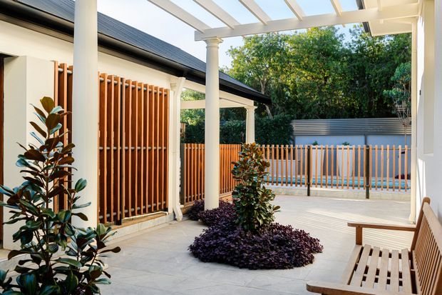 A bright covered outdoor courtyard or terrace with white classical-style columns and pergola beams, pale tiled paving, timber vertical privacy screens, a slatted timber bench, and landscaped planting around a central garden bed. A swimming pool is visible beyond the timber and metal balustrade, with dense greenery providing privacy.